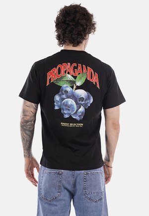 Uomo con tatuaggi indossa una t-shirt nera con mirtilli con facce di teschio, foglie verdi, la scritta "PROPAGANDA" in rosso sopra e un piccolo testo giallo sotto.