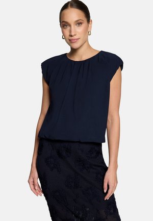 Femme aux cheveux tirés en arrière portant des boucles d'oreilles créoles en or, une blouse sans manches plissée bleu marine et une jupe texturée foncée sur un fond uni.