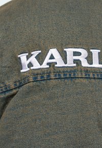 Kurtka dżinsowa w oliwkowym kolorze z białym haftowanym napisem "KARL" z tyłu, wykonana z klasycznej średniej wagi tkaniny z przeszyciami.