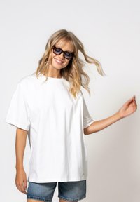 Bílá oversized bavlněná trička s krátkým rukávem, s kulatým výstřihem, v kombinaci s džínovými šortkami, na jednobarevném pozadí.