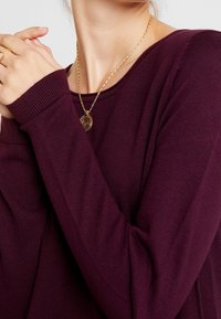 Bordeaux pitkähihaiseen neuleeseen, jossa on ribbireunat ja teksturoitu neulonta. Kaulalla näkyy herkkä kultaketju, jossa on pyöreä riipus.