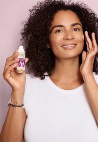 Durchsichtige Kunststoffpumpflasche mit Hautserum und lila Etikett, gehalten von einer Person mit lockigem Haar. Heller lila Hintergrund; glatte Hauttextur.