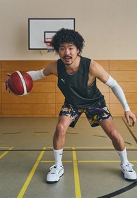 Jugador de baloncesto en una camiseta sin mangas negra y pantalones cortos de colores vibrantes, botando un balón de baloncesto rojo y blanco en un gimnasio.