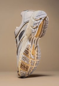 Chaussure de course Reebok beige et blanche inclinée pour montrer la semelle texturée détaillée et la tige en mesh sur un fond beige uni.