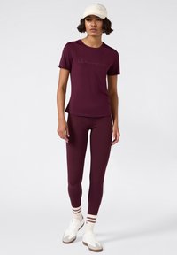Mujer vestida con una camiseta de mangas cortas color burdeos y leggings, zapatillas blancas, calcetas a rayas y una gorra de béisbol beige, de pie frente a un fondo liso.