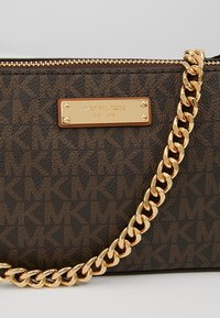 Sac marron Michael Kors avec motif répété du logo MK, plaque dorée indiquant "Michael Kors Est 1981," et une épaisse chaîne dorée comme bandoulière.