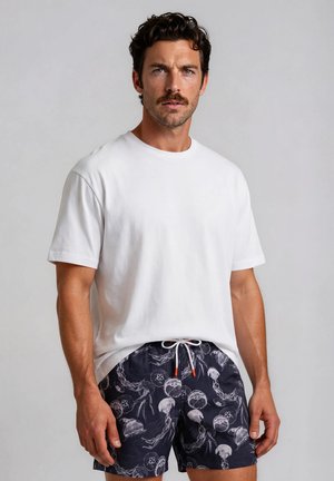 Man with dark curly hair and mustache wearing plain white t-shirt and black swim shorts with white jellyfish pattern, standing against gray background.