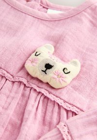 Robe en lin rose ornée d'un patch en forme d'ours texturé avec des fleurs brodées. Détail à volants à la taille et aux manches. Design doux et délicat.