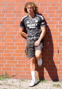 Svart og hvit idrettsjersey med "UNIBET"-tekst, kombinert med grå camouflage cargo-shorts og hvite joggesko, mot en murvegg.