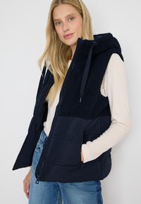 Chaleco azul marino con capucha, que presenta una sección superior de forro polar y una sección inferior acolchada. Bolsillos con cremallera a ambos lados.