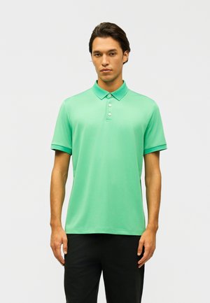 Jonge man staand, gekleed in een mintgroen poloshirt met korte mouwen en zwarte broek, tegen een effen lichte achtergrond.