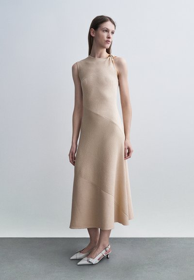Mujer de pie vistiendo un vestido midi beige sin mangas con textura acanalada, escote asimétrico y tacones slingback blancos de punta puntiaguda.
