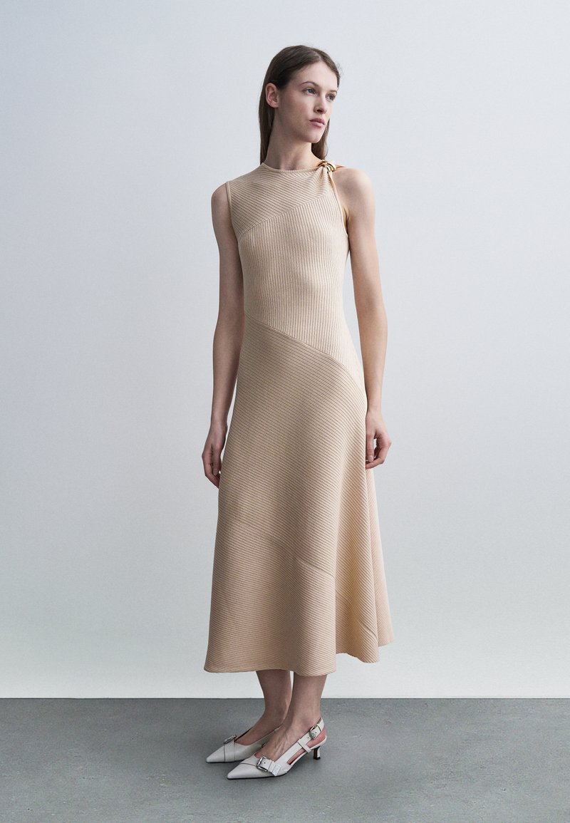 Femme debout portant une robe midi beige sans manches à côtes avec un décolleté asymétrique et des escarpins blancs à bout pointu avec bride arrière.