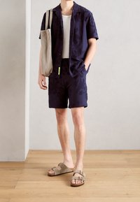 Persona che indossa pantaloni corti floreali blu navy, una camicia a maniche corte abbinata, una canotta bianca, sandali beige e porta una borsa a tracolla beige.