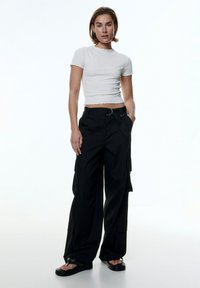 Witte cropped ribbed shirt met golvende randen, gecombineerd met losse zwarte cargobroek met zijzakken en een metalen riemlus. Zwarte sandalen.