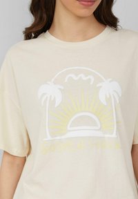 Crème-kleurig t-shirt met een witte en gele grafische afbeelding van een zonsopgang met palmbomen en de tekst "GOLDEN HOUR" eronder.