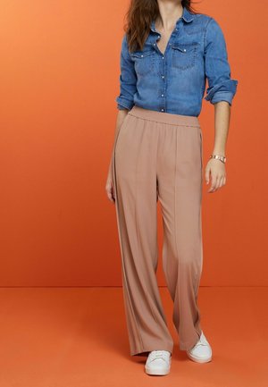 Femme debout devant un fond orange portant une chemise en jean retroussée, un pantalon beige à jambes larges, des baskets blanches et une montre-bracelet.