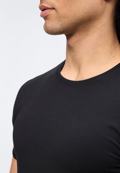 Νεαρό άτομο που φοράει εφαρμοστό μαύρο μπλουζάκι με στρογγυλή λαιμόκοψη, φωτογραφημένο από τον λαιμό έως το στήθος, με φόντο ένα απλό φωτεινό φόντο.