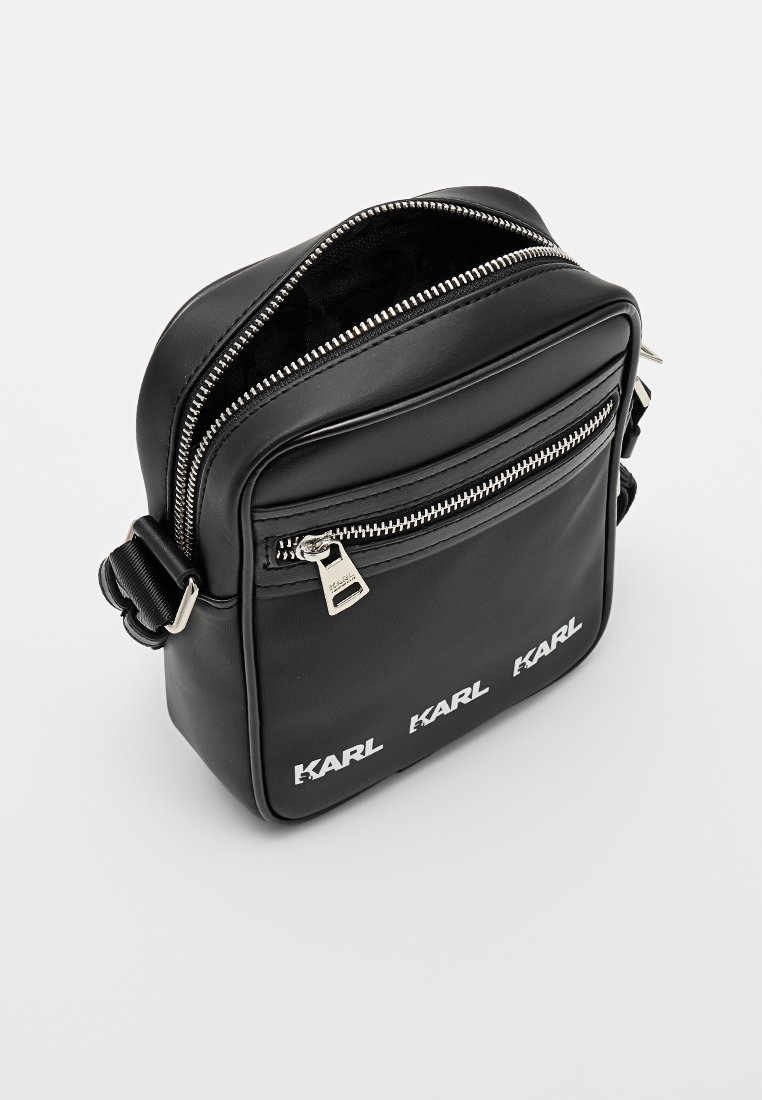 Bolso de hombro sintético negro con cremalleras plateadas, que presenta un bolsillo frontal con cremallera y un logotipo blanco en la parte inferior. Forma cuadrada y compacta.