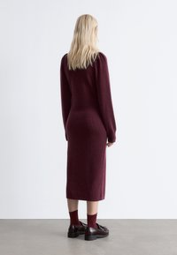 Weinrotes Strickkleid mit langen Ärmeln und gerader Silhouette. Verfügt über gerippte Details an den Bündchen. Getragen mit dunklen Slippern und Socken.