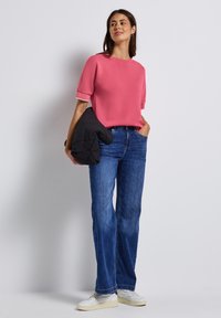 Haut rose à manches courtes avec des accents blancs, associé à un jean bleu à jambes larges et des baskets blanches. Le mannequin tient une veste en doudoune noire.
