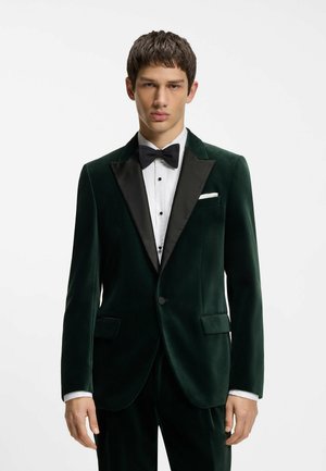 Giovane uomo che indossa una giacca da smoking in velluto verde scuro con revers in raso nero, camicia bianca, papillon nero e fazzoletto bianco da taschino.