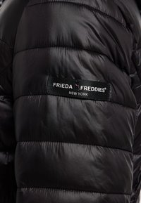 Zwarte pufferjack met een gladde, glanzende stof en horizontale stiknaden. Inclusief een zwart label met de tekst "FRIEDA FREDDIES® NEW YORK."