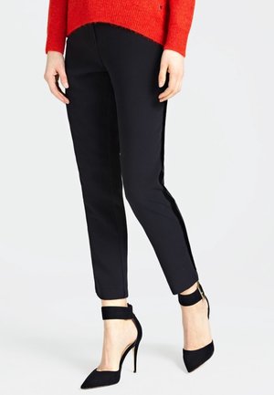 Kobieta w czarnych wąskich spodniach do kostek, czarnych szpilkach z paskiem wokół kostki oraz czerwonej bluzce z długim rękawem, stojąca na jednolitym tle.