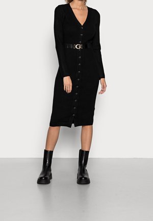 Kobieta ubrana w czarną midi sukienkę na guziki z długimi rękawami oraz szerokim czarnym paskiem z złotą klamrą, w połączeniu z czarnymi botkami.