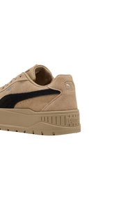 Zapatilla de ante beige con acentos de diseño negro, logo en el lateral y suela de goma texturizada con surcos. Estilo de plataforma elevada.
