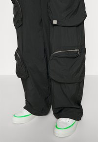 Zwarte, lichtgewicht jumpsuit van stof met meerdere ritszakken; gecombineerd met witte sneakers met groene accenten op de zool en de tong.