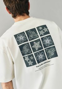 Hvid t-shirt med korte ærmer, der har et mørkt grafik af ni blomsterdesigns på ryggen, ledsaget af tekst nederst.