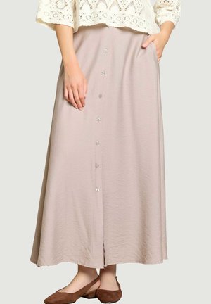 Femme portant une jupe midi beige boutonnée sur le devant, une main dans la poche et des chaussures plates slingback marron, associée à un top en dentelle blanc.