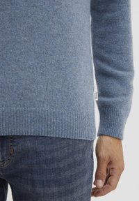 Lys blå genser synlig ved kanten og mansjettene, laget av strikket stoff med ribbestruktur, kombinert med mørke jeans.