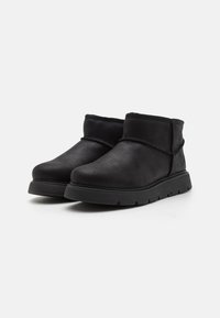 Bottines noires en daim doux avec un bout arrondi, une semelle en caoutchouc basse et un détail de poignet élastique. Texture lisse avec un minimum de coutures.