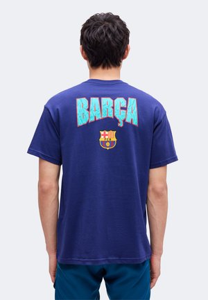 Personne portant un t-shirt bleu avec un grand texte turquoise "BARÇA" et un écusson du club de football de Barcelone dans le dos, debout de dos.