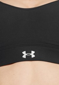 Schwarzer Sport-BH aus strukturiertem Stoff, mit einem breiten Bund und einem weißen Under Armour-Logo in der Mitte. Glatte, abgerundete Ausschnittform.