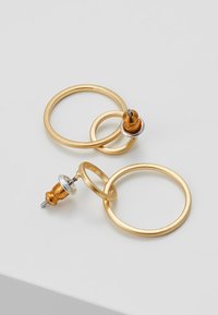 Dvoje zlatých kovových náušnic ve tvaru smyčky spojených dohromady s pérovým uzávěrem na světle šedém povrchu.