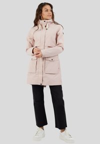 Parka imperméable rose avec un col haut, fermeture éclair, deux grandes poches avant et poignets ajustables ; porté sur un pantalon noir et des baskets blanches.