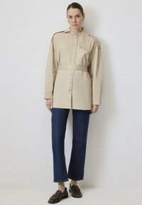 Camisa chaqueta grande beige con botones en la parte delantera y cintura ajustada, combinada con jeans rectos de color azul oscuro y mocasines oscuros.