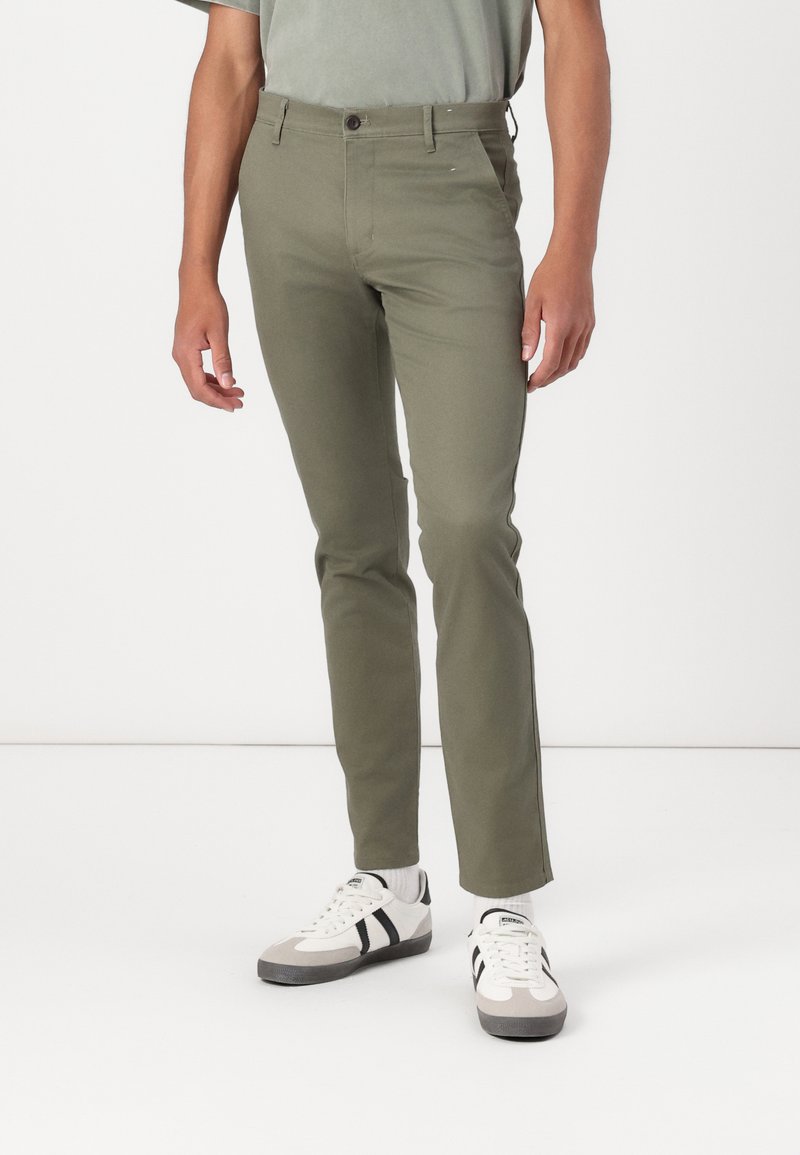 Pantalons fuselés vert olive avec une texture lisse, fermeture à bouton et poches latérales, associés à des baskets blanches avec des accents noirs.
