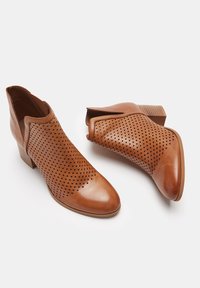 Brązowe skórzane botki z perforowanym wzorem, spiczastym noskiem i obcasem warstwowym. Wierzch ma gładkie wykończenie i unikalny wycięcie dla dodatkowego detalu.