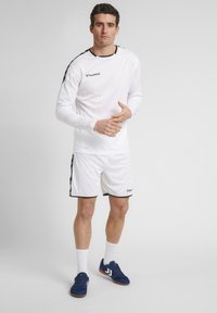 Hvid langærmet træningsskjorte med sorte detaljer, parret med hvide shorts med sorte kanter. Båret med blå træningssko og hvide sokker.