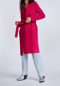 Fuksiansävyinen takki, jossa on kaulus, pitkät hihat ja vyötärösolki. Yhdistetty vaaleansinisiin kulutuspintaisiin farkkuihin ja mustavalkokuvioituihin nilkkureihin.