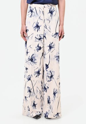 Femme portant un pantalon large taille haute crème avec un imprimé floral noir et bleu marine, associé à un haut foncé et des bagues aux deux mains.