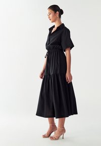 Vestito nero a camicia con maniche corte, vita cinchata, gonna a strati e cordoncino. Abbinato a tacchi leggeri con cinturini. Tessuto morbido.