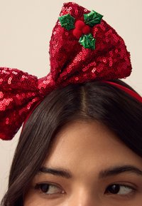 Rotes Haarband mit Pailletten und großer Schleife, geschmückt mit grünen Stechpalmenblättern und roten Bommel in der Mitte. Glatte Textur mit glänzenden Pailletten.