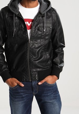 Homme portant une veste en cuir noire avec une capuche grise et un t-shirt blanc avec un logo rouge, mains dans les poches, jean bleu visible de la taille vers le bas.