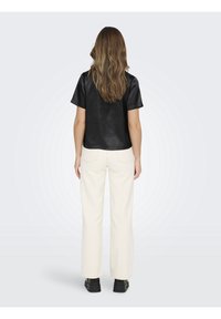 Vrouw met lang golvend haar, gekleed in een zwart shirt met korte mouwen, witte rechte broek en zwarte chunky schoenen, staand met haar rug naar de kijker gekeerd.