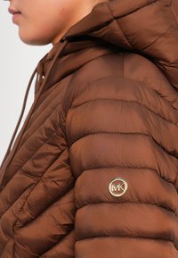 Sidelengs av en person som har på seg en brun, quiltet hettejakke med et rundt, gyllent MK-logo på den øvre ermen.